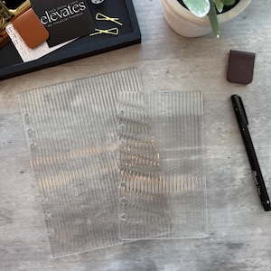 May include: Clear, vertically-lined plastic binder inserts with punched holes. A black tray holds a business card with the text "elevates my financial freedom", a dice, and gold paper clips. A brown leather bookmark and a black Sharpie pen are also present.