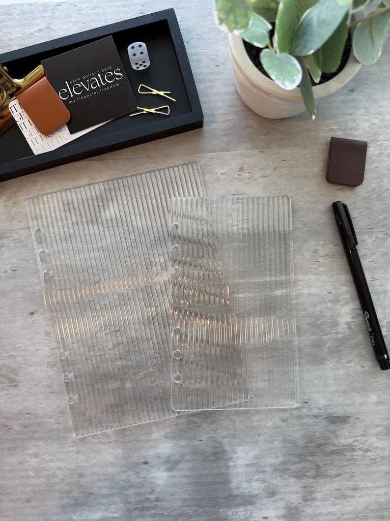 May include: Clear, vertically-lined plastic binder inserts with punched holes. A black tray holds a business card with the text "elevates my financial freedom", a dice, and gold paper clips. A brown leather bookmark and a black Sharpie pen are also present.