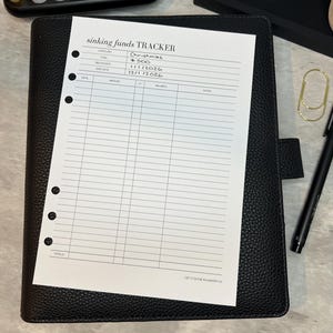 May include: A black leather planner with a "sinking funds TRACKER" sheet. The sheet includes sections for category, goal, begin date, end date, date, amount, balance, and notes. A calculator and pen are also visible.