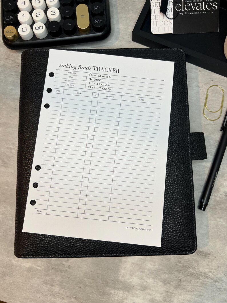 May include: A black leather planner with a "sinking funds TRACKER" sheet. The sheet includes sections for category, goal, begin date, end date, date, amount, balance, and notes. A calculator and pen are also visible.