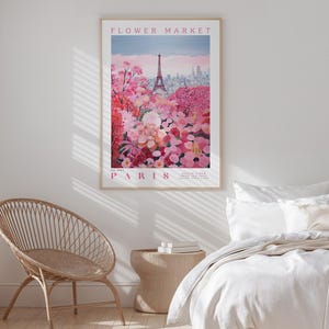 a bedroom with a large poster of a flower market in Paris hanging on the wall. The poster depicts a vibrant scene of pink and red flowers, with the Eiffel Tower visible in the background. The bedroom has a bed with white linens and a wicker chair, creating a cozy and inviting atmosphere.