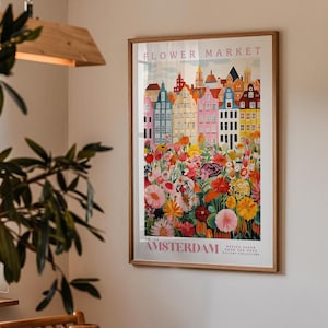 a framed poster of a colorful cityscape with flowers, hanging on a wall in a room with a plant in the foreground.