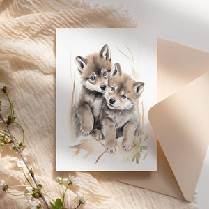 Op de afbeelding: Een aquarel schilderij van twee wolf pups die tegen elkaar aan knuffelen. De pups zijn bruin en wit met blauwe ogen. De achtergrond is een lichte waslaag in groen en bruin.