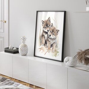 Op de afbeelding: Een aquarel schilderij van twee wolf pups die knuffelen. De pups zijn bruin en wit en hebben zwarte neuzen. Het schilderij is ingelijst in een zwart lijst en hangt aan een witte muur.