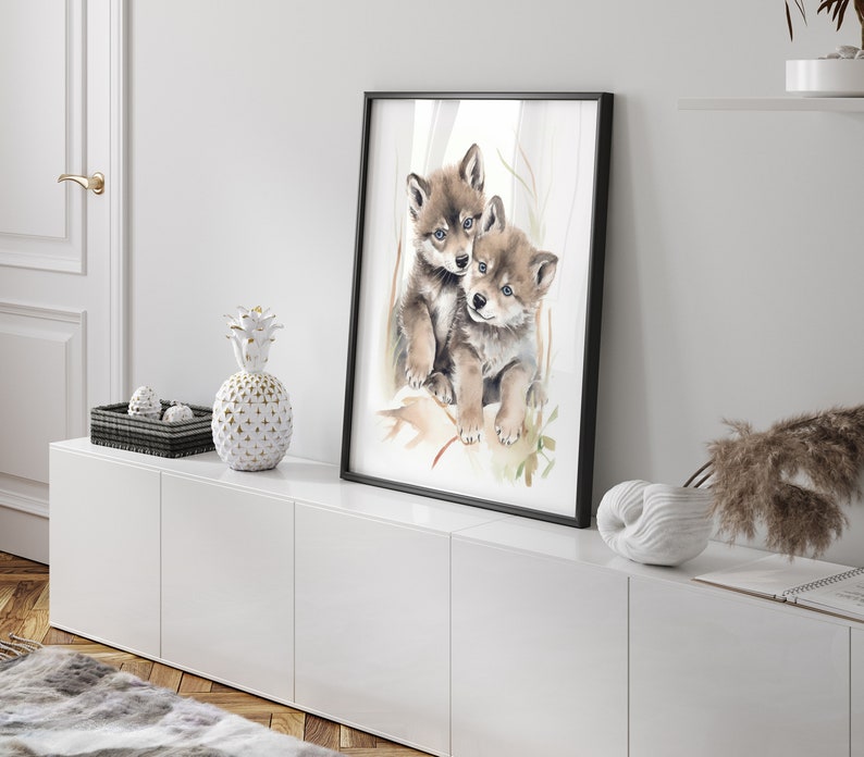 Op de afbeelding: Een aquarel schilderij van twee wolf pups die knuffelen. De pups zijn bruin en wit en hebben zwarte neuzen. Het schilderij is ingelijst in een zwart lijst en hangt aan een witte muur.
