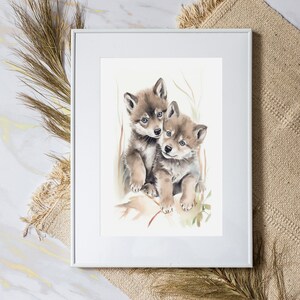 Op de afbeelding: Een aquarel schilderij van twee wolf pups die knuffelen. De pups zijn bruin en grijs met witte markeringen. Het schilderij is tegen een witte achtergrond met een lichtbruine lijst geplaatst.