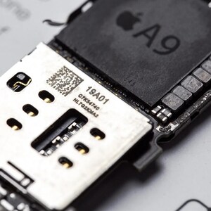 Op de afbeelding: Een zwarte Apple A9 processorchip gemonteerd op een printplaat. De plaat bevat een zilverkleurig metalen onderdeel met een QR-code en de gedrukte tekst 19A01, C7K54740 en HL7G2KMA2. Goudkleurige contactpunten zijn zichtbaar op het onderdeel.