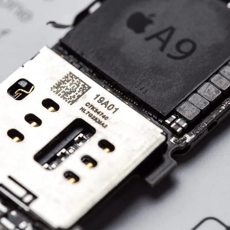 Op de afbeelding: Een zwarte Apple A9 processorchip gemonteerd op een printplaat. De plaat bevat een zilverkleurig metalen onderdeel met een QR-code en de gedrukte tekst 19A01, C7K54740 en HL7G2KMA2. Goudkleurige contactpunten zijn zichtbaar op het onderdeel.