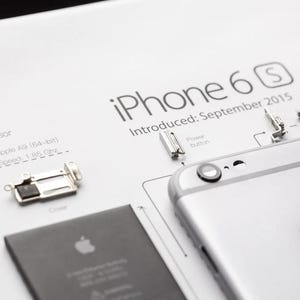 Op de afbeelding: Een wit technisch diagram van een iPhone 6s met de tekst "iPhone 6s" en "Introduced: September 2015". De afbeelding toont een zilveren smartphone, een zwarte batterij en kleine interne metalen onderdelen met het label "Processor" en "Power button".