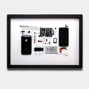 May include: A black framed display featuring a disassembled iPhone 3G with its internal components, including the logic board, battery, and screen, laid out on a white background with technical labels and text describing the device's history.