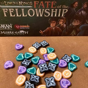Può includere: È presente una scatola di gioco da tavolo con il titolo "The Lord of the Rings: Fate of the Fellowship". Pezzi di gioco colorati in varie forme e colori, tra cui viola, arancione, verde acqua e nero, sono sparsi davanti alla scatola.