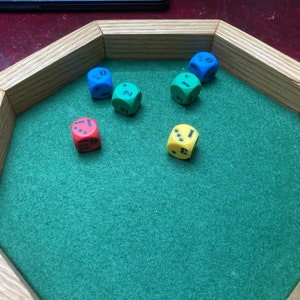 May include: A wooden dice tray with a green felt interior holds six multi-colored dice. The dice are blue, green, red, and yellow, each showing different numbers. The tray has an octagonal shape.