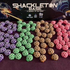 May include: A set of 50 hexagonal game pieces in five different colours: purple, green, brown, pink, and white. The pieces are arranged in groups of 10, with each group forming a star shape. The game pieces are made of plastic and have a honeycomb pattern.