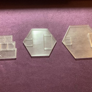 Può includere: Tre espositori in plastica trasparente e geometrica. Uno ha una forma rettangolare a gradini, e gli altri due sono esagonali con ritagli rettangolari. Gli espositori sono su una superficie viola.