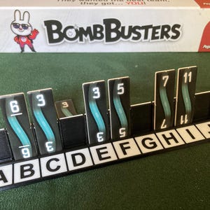 以下が含まれることがあります： 「Bomb Busters」というボードゲーム。ゲームの駒と箱があります。駒は黒色で、白い数字とターコイズのデザインが施されています。箱には漫画のウサギとPegasus Spieleのロゴが描かれています。