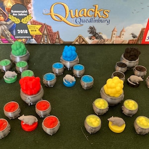 May include: A game board with the title "The Quacks of Quedlinburg" and a variety of colorful game pieces, including green, blue, yellow, and red. The game pieces are arranged in a circle around the board, with a small, gray mouse figurine in the center of each circle.