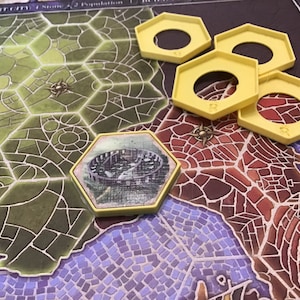 Puede incluir: Una pieza de juego hexagonal amarilla con un agujero en el centro. La pieza está en un tablero de juego con un patrón verde y marrón. La pieza está al lado de una pieza hexagonal más pequeña con una imagen de un edificio en ella.