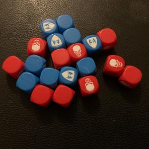 May include: A collection of red and blue dice with white symbols. The dice are scattered on a dark surface. The symbols include a skull, a shield, and a bearded face. These dice are likely used for tabletop gaming.