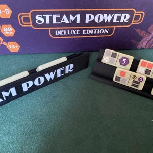 Puede incluir: El juego de mesa "Steam Power Deluxe Edition" se muestra con piezas y la caja. La caja es morada con el título del juego en blanco y naranja. Las piezas son beige con gráficos y números coloridos.