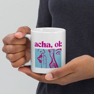 May include: White ceramic mug with a pink and blue illustration of two people whispering. The text "acha, ok" is printed in pink on the mug.