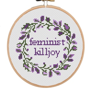 May include: A cross-stitch embroidery hoop with the words "feminist killjoy" in purple, encircled by a wreath of green leaves and purple flowers. The design is set against a white background.