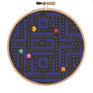 May include: A cross-stitch embroidery hoop featuring a Pac-Man game design. The design includes a maze in shades of blue and black, with colorful Pac-Man characters. The wooden hoop is round and has a brass adjustment screw.