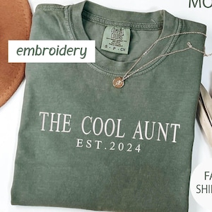 May include: A moss green t-shirt with the text "The Cool Aunt Est. 2024" embroidered in white on the front.