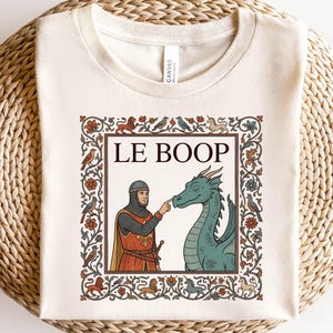 May include: A cream-colored t-shirt featuring a medieval-style graphic print. The design shows a knight in chainmail touching the nose of a green dragon, with the text LE BOOP above them, all enclosed in a decorative floral and animal border.