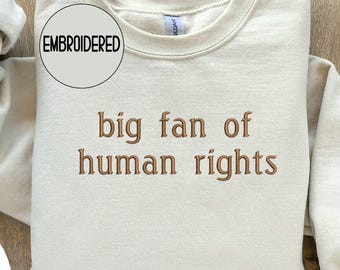 May include: A cream-colored sweatshirt with the embroidered phrase "big fan of human rights" in brown thread. The sweatshirt has a crew neck and long sleeves. The word "EMBROIDERED" is in a circle above the text.