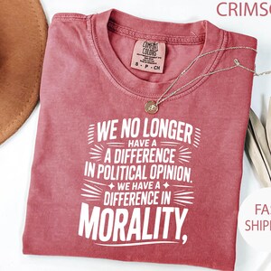 May include: Crimson-colored t-shirt with the text "WE NO LONGER HAVE A DIFFERENCE IN POLITICAL OPINION. WE HAVE A DIFFERENCE IN MORALITY." A gold necklace rests on the shirt. The shirt is labeled "Comfort Colors" and is size S-P-CH.