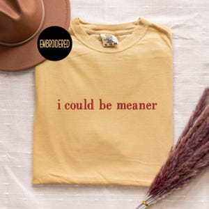 May include: Mustard yellow t-shirt with the phrase "i could be meaner" in red lettering. The shirt is folded and displayed on a white surface. A brown hat and a bundle of dried flowers are also in the image. A black circle with the word "EMBROIDERED" is in the upper left corner.