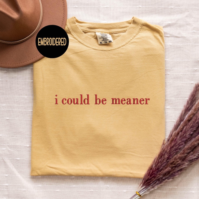 May include: Mustard yellow t-shirt with the phrase "i could be meaner" in red lettering. The shirt is folded and displayed on a white surface. A brown hat and a bundle of dried flowers are also in the image. A black circle with the word "EMBROIDERED" is in the upper left corner.
