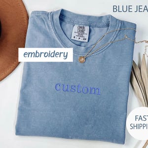May include: A blue jean t-shirt with the word "custom" embroidered in blue thread. The shirt is folded and laid flat on a white surface.