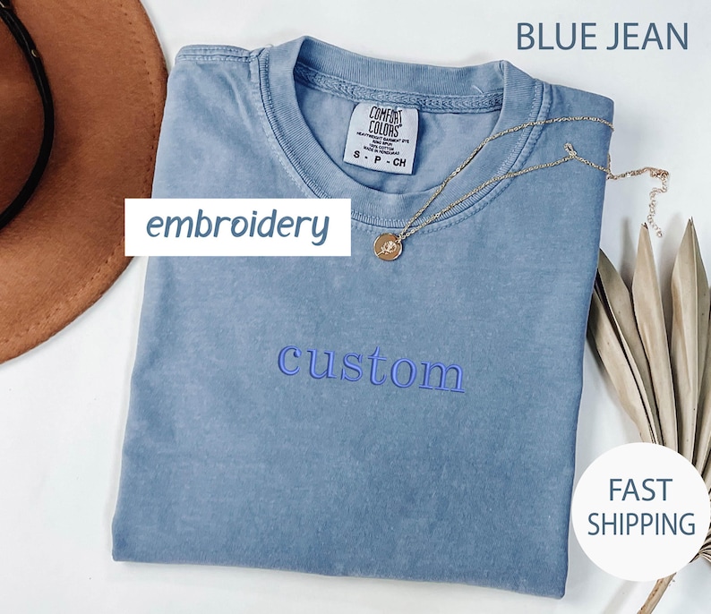 May include: A blue jean t-shirt with the word "custom" embroidered in blue thread. The shirt is folded and laid flat on a white surface.