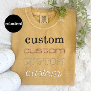 May include: A mustard yellow t-shirt with the word "custom" embroidered in four different styles: black, pink, white, and a white outline.