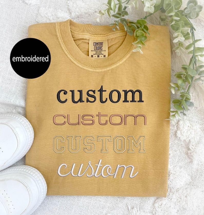May include: A mustard yellow t-shirt with the word "custom" embroidered in four different styles: black, pink, white, and a white outline.