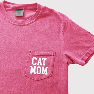 May include: A pink t-shirt with a pocket that reads "CAT MOM" in white block letters. The shirt has a crew neck and short sleeves. The tag in the collar reads "COMFORT COLORS".