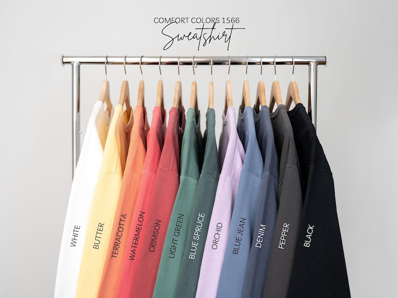 May include: A rack of 12 sweatshirts in various colors, including white, butter, terracotta, watermelon, crimson, light green, blue spruce, orchid, blue jean, denim, pepper, and black. The sweatshirts are labeled with the color names below each one. The text "COMFORT COLORS 1566 Sweatshirt" is at the top of the image.