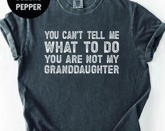 May include: Dark gray t-shirt with the text "YOU CAN'T TELL ME WHAT TO DO YOU ARE NOT MY GRANDDAUGHTER" in white. The shirt also has the words "COMFORT COLORS PEPPER" in a black circle. The shirt is a crew neck.