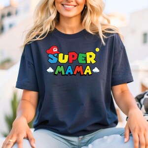 May include: A navy blue t-shirt with a colorful graphic of a Super Mario-inspired design. The graphic features the text "SUPER MAMA" with a red and white Mario hat.