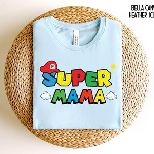May include: Light blue t-shirt with a graphic of a Super Mario character wearing a red cap and the text "SUPER MAMA" in colorful letters.