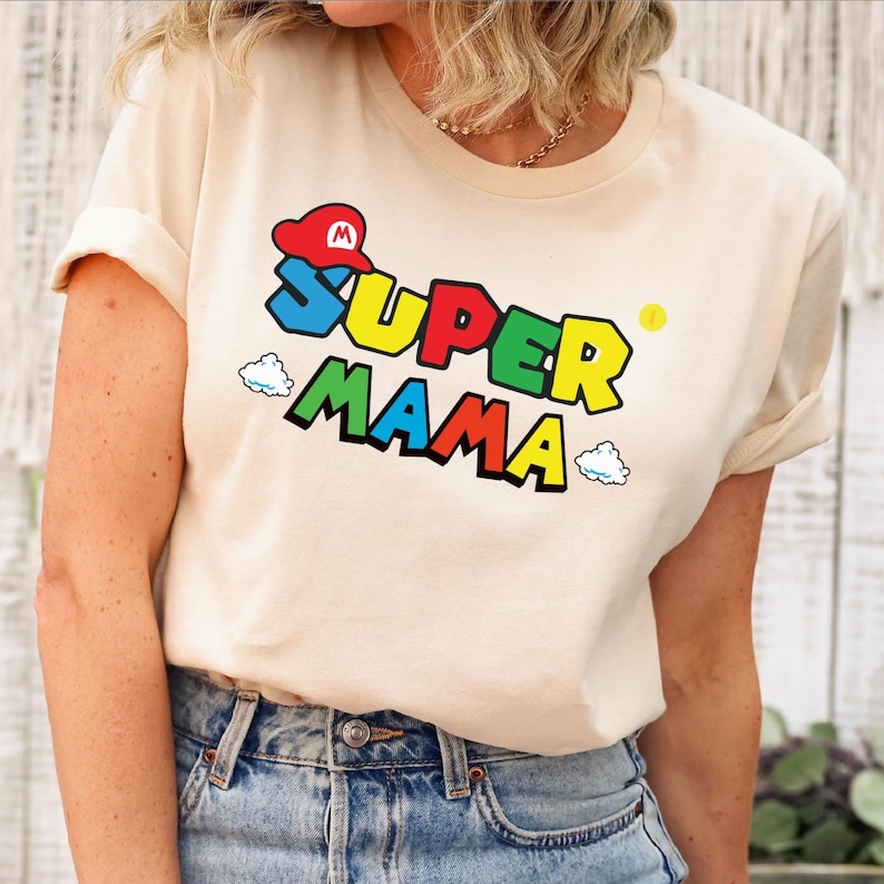 May include: A beige t-shirt with a colorful graphic that says "SUPER MAMA" with a red and white Mario hat above the text.