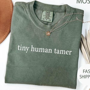May include: An olive green t-shirt with the text "tiny human tamer" in white. A gold-colored necklace with a small pendant rests on the shirt. The shirt has a crew neck and short sleeves. The tag reads "Comfort Colors".