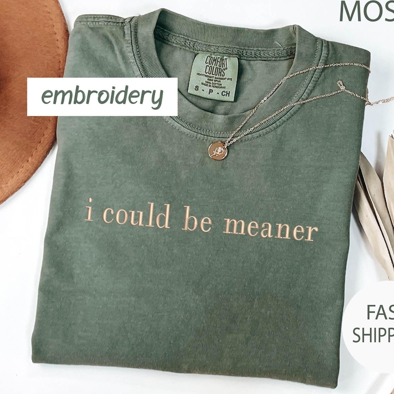May include: Olive green t-shirt with the embroidered phrase "i could be meaner" in a light brown color. A gold-colored necklace with a pendant rests on the shirt. The shirt has a Comfort Colors tag.