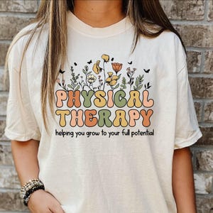 May include: Cream-colored t-shirt with the words "PHYSICAL THERAPY" in retro-style lettering, accompanied by a floral design. The shirt also includes the text "helping you grow to your full potential".