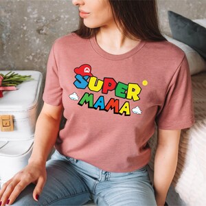 May include: A pink t-shirt with a colorful graphic of a Super Mario-inspired character wearing a red hat and the text "SUPER MAMA".