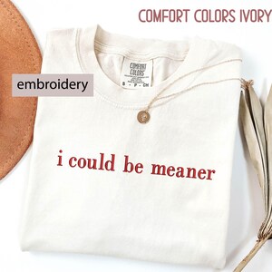 May include: Ivory-colored t-shirt with the embroidered phrase "i could be meaner" in red. The shirt is from Comfort Colors and has a gold necklace draped across it. A brown hat and dried plant are in the background.