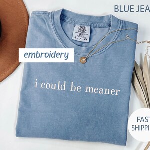 May include: A blue jean colored t-shirt with the embroidered phrase "i could be meaner". The shirt has a crew neck and short sleeves. A gold necklace with a pendant is draped on the shirt. The word "embroidery" is in a white box.