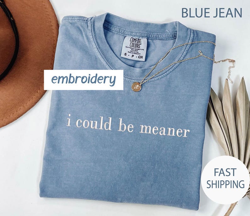 May include: A blue jean colored t-shirt with the embroidered phrase "i could be meaner". The shirt has a crew neck and short sleeves. A gold necklace with a pendant is draped on the shirt. The word "embroidery" is in a white box.