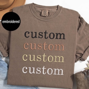 May include: A brown t-shirt with the word "custom" embroidered on it in four different colors: black, peach, yellow, and white.